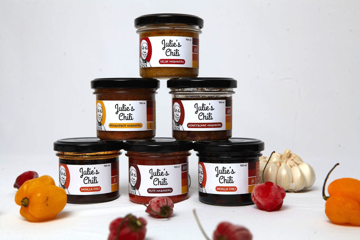 Five jars of Julio's Chili hot sauce arranged in a pyramid with whole peppers, garlic, and chili peppers placed around them on a white background.