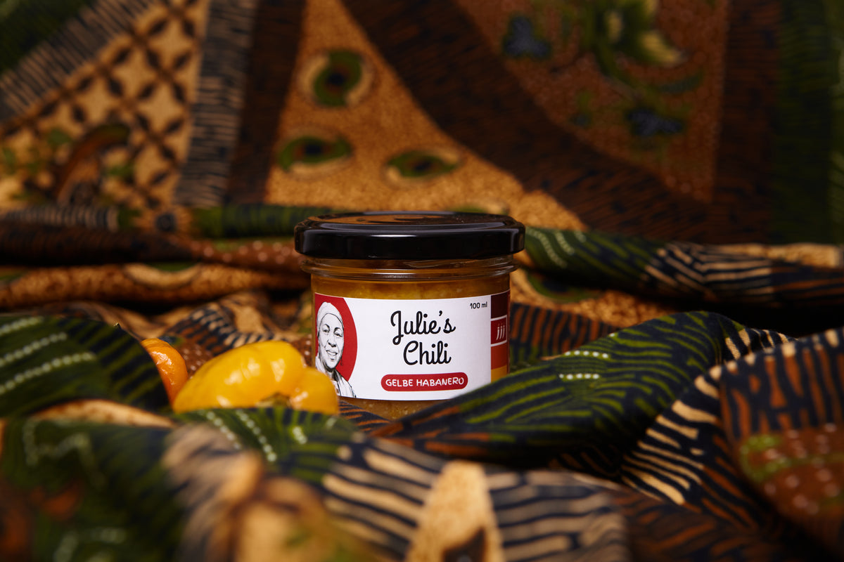 A jar of Julie's Chili Guelfe Habanero sauce sits on a patterned green and brown textile with blurred yellow habanero peppers in the foreground.